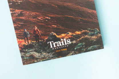The bottom half of a magazine is over a blue background. The cover image is of a mountain ridge that is illuminated in reds and oranges from the sunrise. There are wo people, one earing a blue jacket and the othe rin a maroon jacket, hiking uphill toward the photographer. The text "Trails Magazine - Issue Three" is centered at the bottom. In the top right corner is the logo