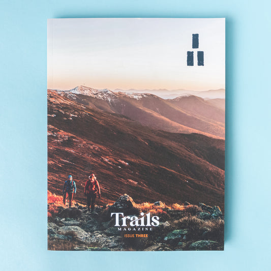 A magazine is over a light blue background. The cover image is of a mountain ridge that is illuminated in reds and oranges from the sunrise. There are wo people, one earing a blue jacket and the othe rin a maroon jacket, hiking uphill toward the photographer. The text "Trails Magazine - Issue Three" is centered at the bottom. In the top right corner is the logo, a set of three thick horizontal lines with two on the bottom and one on top to make a triangle. 