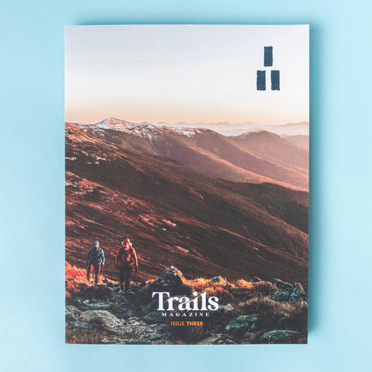 A magazine is over a light blue background. The cover image is of a mountain ridge that is illuminated in reds and oranges from the sunrise. There are wo people, one earing a blue jacket and the othe rin a maroon jacket, hiking uphill toward the photographer. The text "Trails Magazine - Issue Three" is centered at the bottom. In the top right corner is the logo, a set of three thick horizontal lines with two on the bottom and one on top to make a triangle.