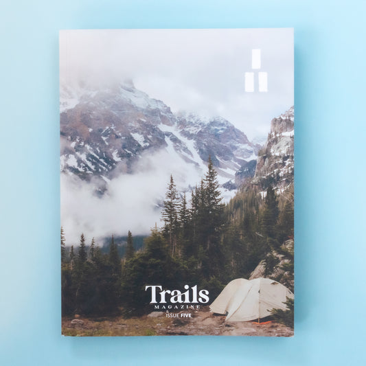 A magazine is over a light blue background. The cover image is of white tent among green pine trees in the backcountry of a cold and foggy mountain scene. Mountian peaks are peeking out of the fog. The text "Trails Magazine - Issue 5" is centered at the bottom. In the top right corner is the logo, a set of three thick horizontal lines with two on the bottom and one on top to make a triangle. 