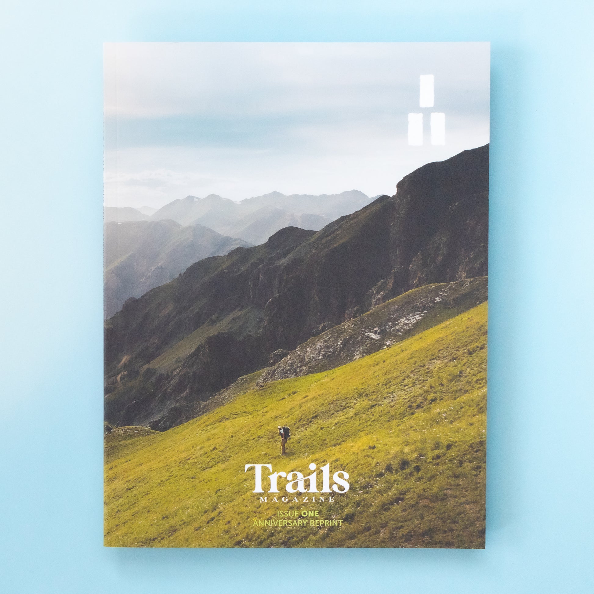 A magazine is over a light blue background. The cover image is of a lush green mountain side with faded peaks off in the distance. In the middle is a person wearing hiking clothes and a backpack hiking downhill. The text "Trails Magazine - Issue One - Anniversary Reprint" is centered at the bottom. In the top right corner is the logo, a set of three thick horizontal lines with two on the bottom and one on top to make a triangle. 