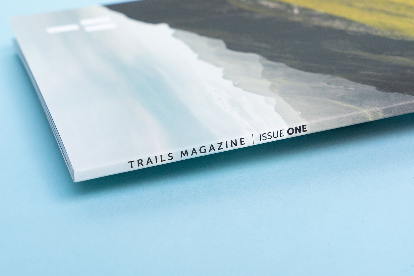 The binding of a magazine, displaying a mountain scene lays across a light blue backdrop. The text along the binding reads, "Trails Magazine Issue One"