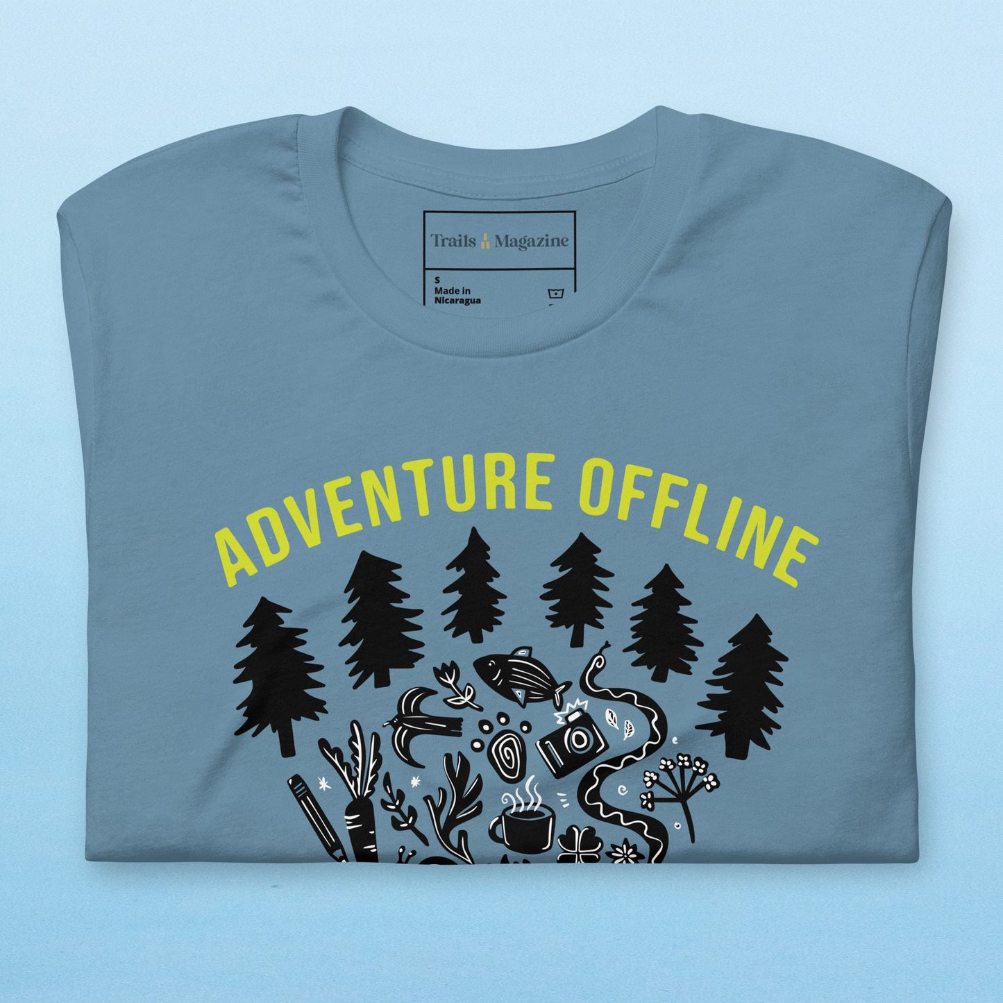 A blue T-shirt is folded over a light blue backdrop. in a neon green, the text "Adventure Offline" arches over a black illustration of pine trees and a collection of items including carrots, flowers, fish, a camera, a snake, coffee cup, and paw print are bundled together. 