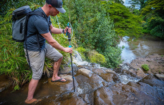 A hiker using the PurTrek water filter trekking pole to drink from a mountain stream.