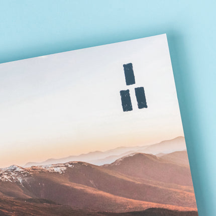 The top right corner of a magazine displaying a mountain ridge that is illuminated in reds and oranges from the sunrise and the Trails Magazine logo, three parallel dark blue rectangles stacked into a triangle making a trail market blaze.