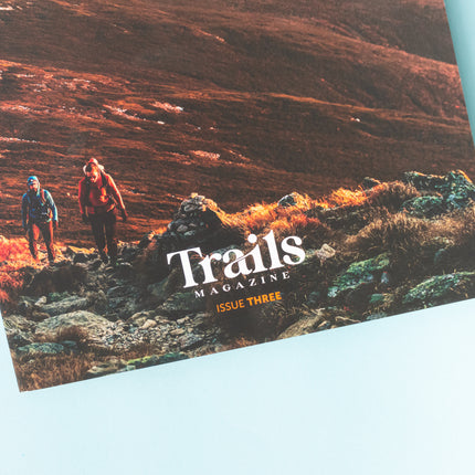 The bottom half of a magazine is over a blue background. The cover image is of a mountain ridge that is illuminated in reds and oranges from the sunrise. There are wo people, one earing a blue jacket and the othe rin a maroon jacket, hiking uphill toward the photographer. The text "Trails Magazine - Issue Three" is centered at the bottom. In the top right corner is the logo