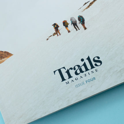 The bottom half of a magazine is over a blue background. The cover displays four women in hiking gear off in the distance, following one another on a hike. The text "Trails Magazine - Issue Four" is centered at the bottom.