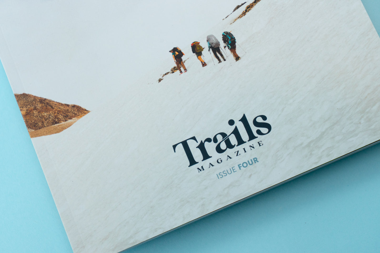 The bottom half of a magazine is over a blue background. The cover displays four women in hiking gear off in the distance, following one another on a hike. The text "Trails Magazine - Issue Four" is centered at the bottom.