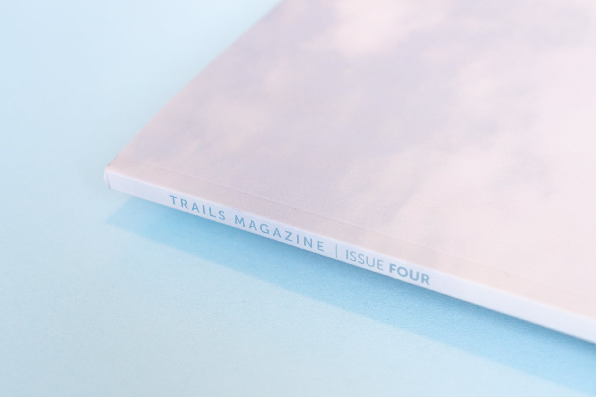 The binding of a magazine, displaying a bright and cloudy sky lays across a light blue backdrop. The text along the binding reads, "Trails Magazine Issue Four" in light blue text.