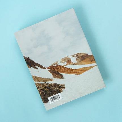 The back of a magazine is over a blue background. The image is a bright snowy mountain scene with peeks of muted yellow and orange tone rocks peeking out of the snow covered ground. 