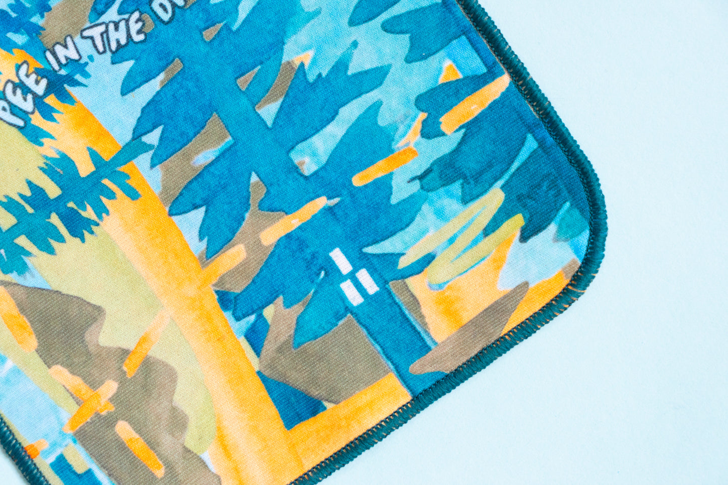 The bottom corner of the Trails and Kula cloth collaboration is visible. The square cloth is painted with an abstract landscape of trees and light in various shades of blue and yellow. Along sone side, the words "Pee in the Dirt." are written. 