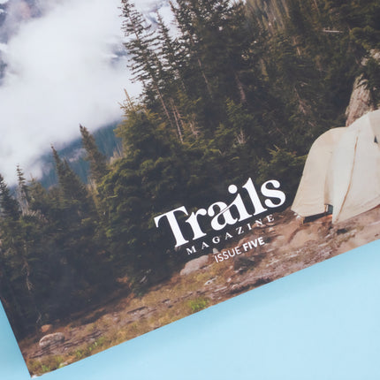 The bottom half of a magazine is over a blue background. The cover image is of white tent in the backcountry of a cold and foggy mountain scene. The text "Trails Magazine - Issue 5" is centered at the bottom.