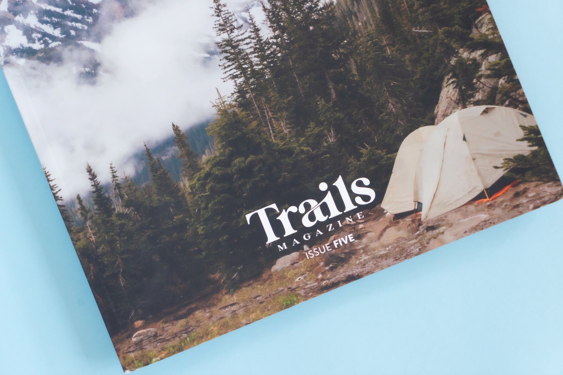 The bottom half of a magazine is over a blue background. The cover image is of white tent in the backcountry of a cold and foggy mountain scene. The text "Trails Magazine - Issue 5" is centered at the bottom.