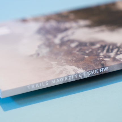 The binding of a magazine, displaying a mountain scene lays across a light blue backdrop. The text along the binding reads, "Trails Magazine Issue 5"