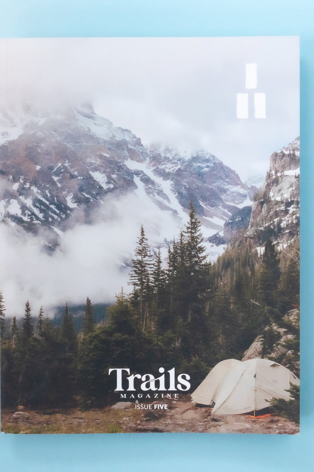 A magazine is over a light blue background. The cover image is of white tent among green pine trees in the backcountry of a cold and foggy mountain scene. Mountian peaks are peeking out of the fog. The text "Trails Magazine - Issue 5" is centered at the bottom. In the top right corner is the logo, a set of three thick horizontal lines with two on the bottom and one on top to make a triangle. 